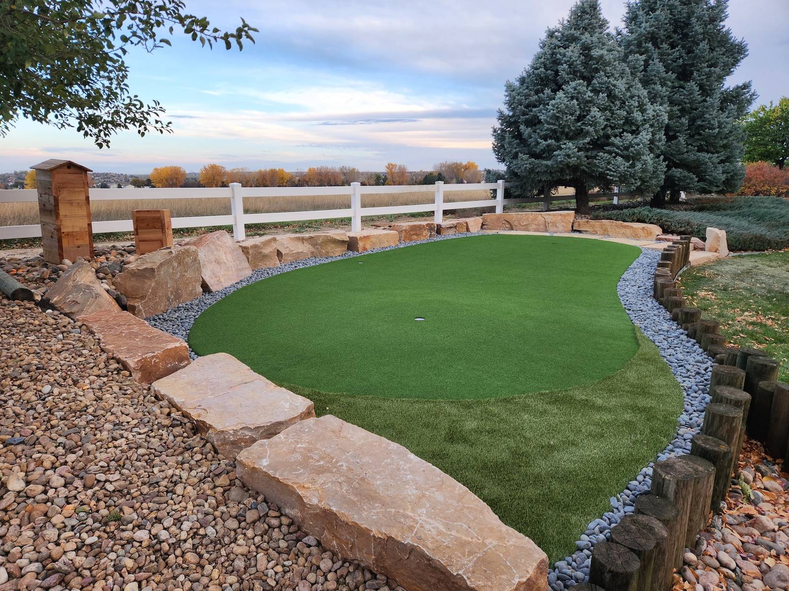 Backyard putting green edged with sandstone boulders, river rock, and cedar posts at sunset