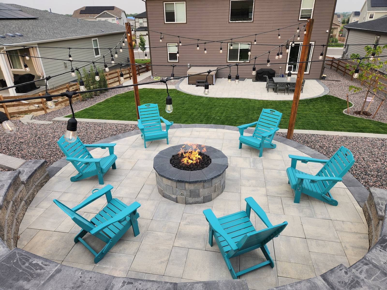 Circular paver fire pit patio with Adirondack seating, turf lawn, and string lighting at dusk
