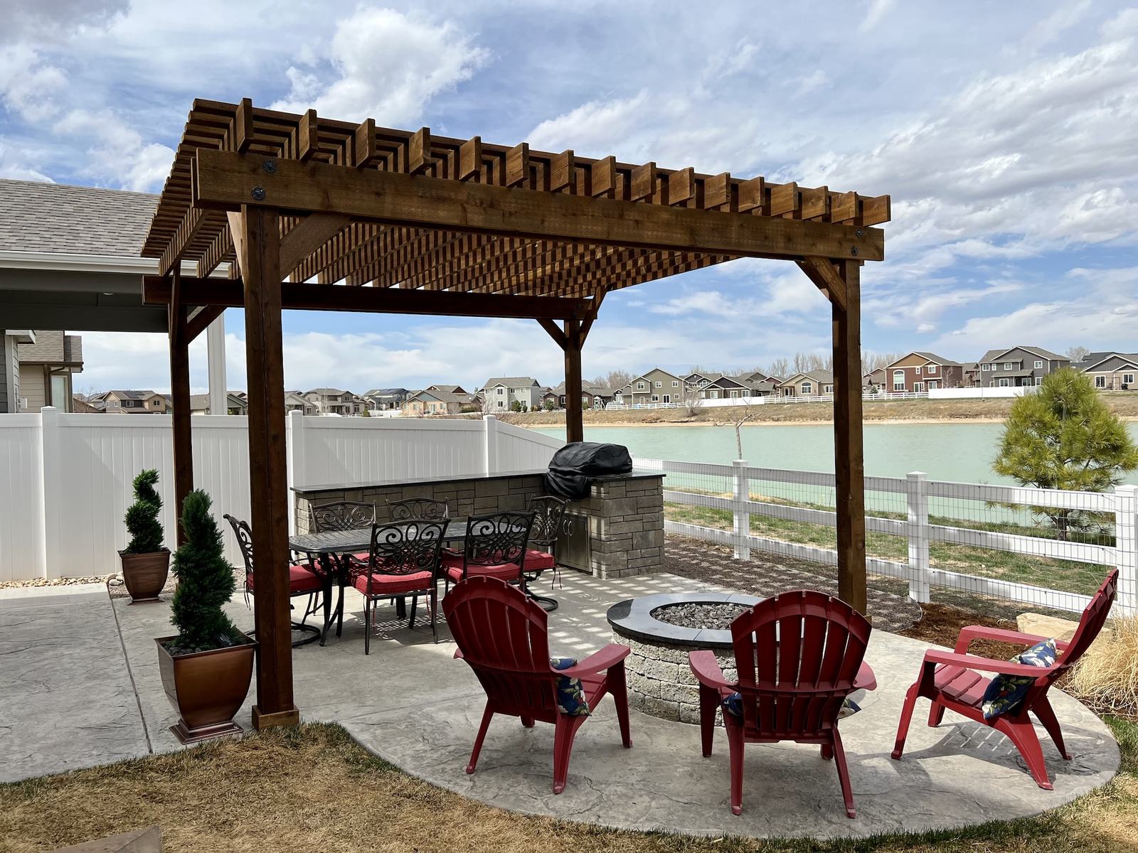 Cedar pergola over a stamped concrete patio with an outdoor kitchen, fire pit, and lake view