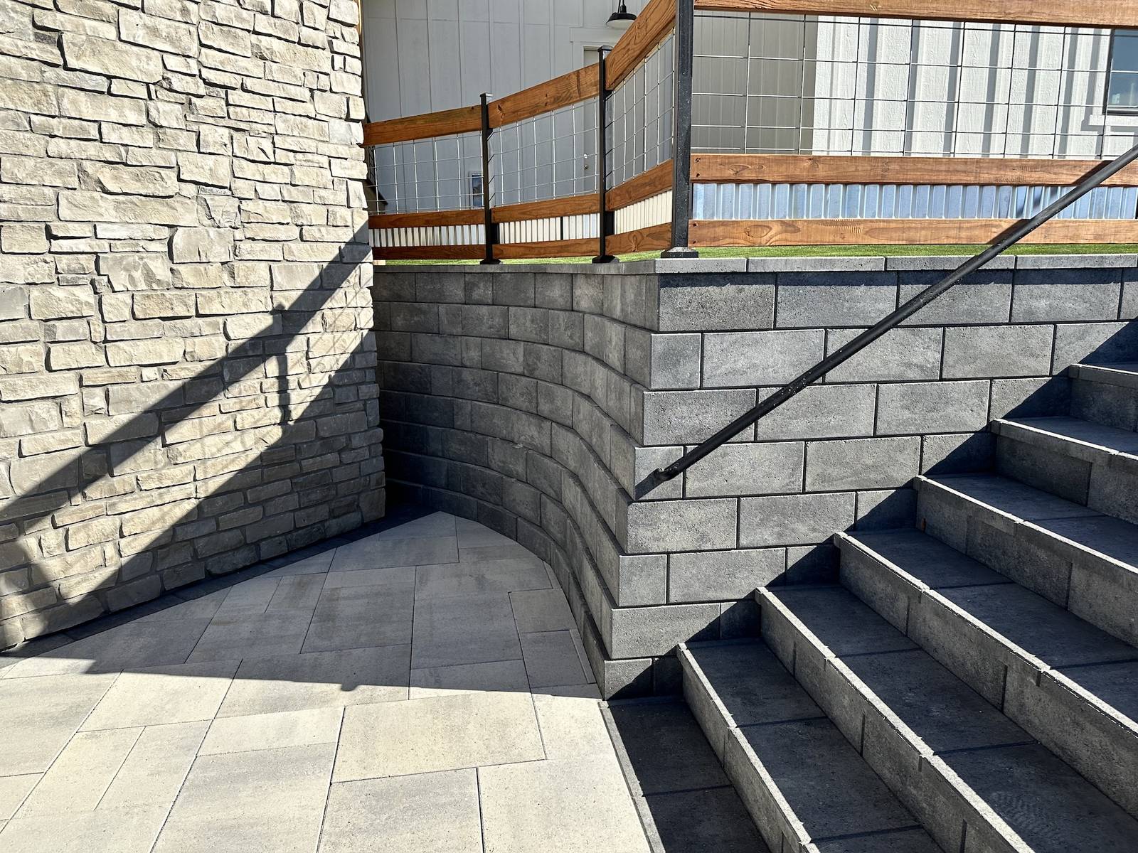 Curved block retaining wall wrapping a paver staircase with a black handrail